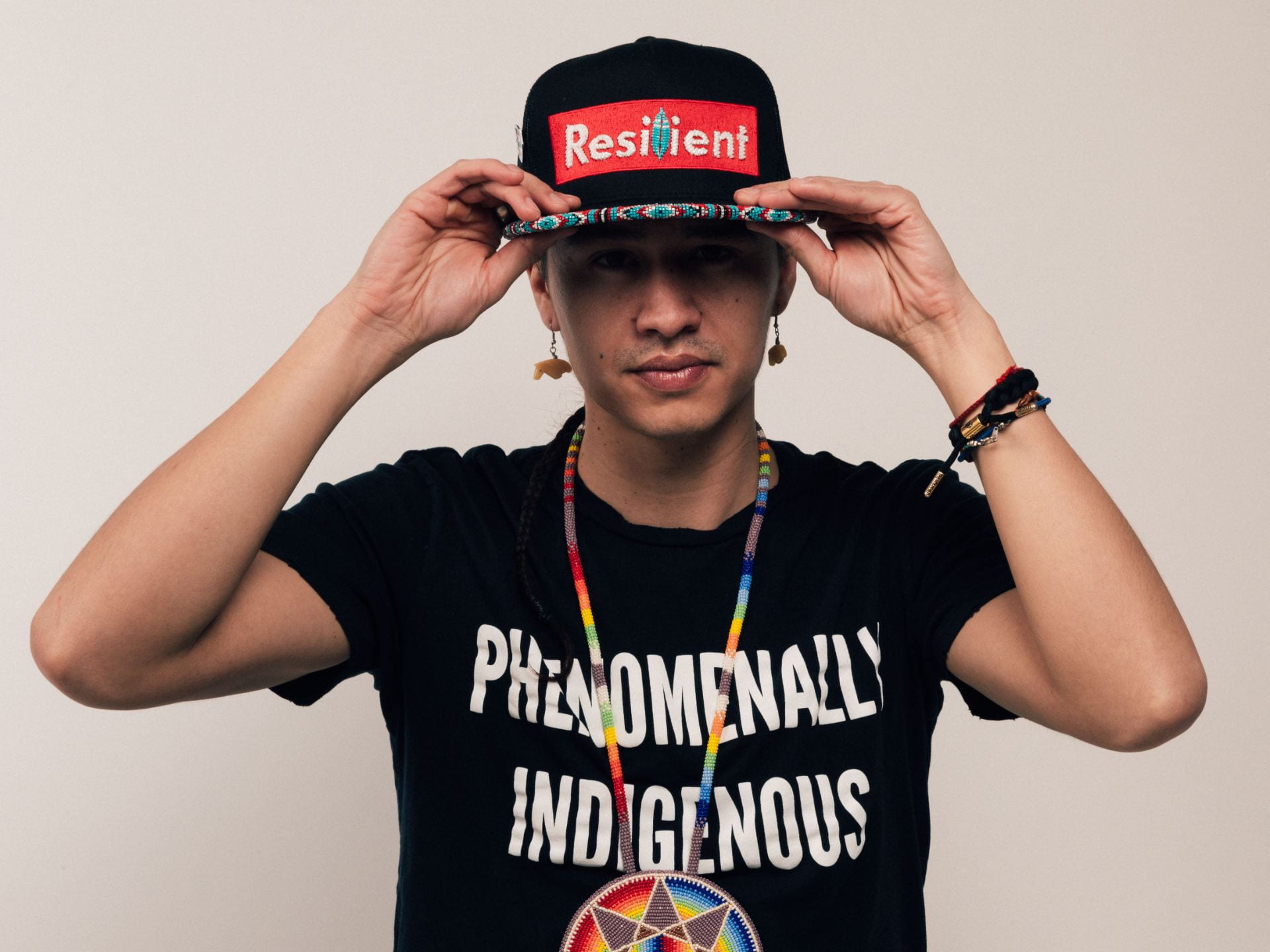 a photo of Frank Waln wearing a cap that says "Resist" in the "Supreme" style, and a t-shirt that says "Phenomenally Indigenous"